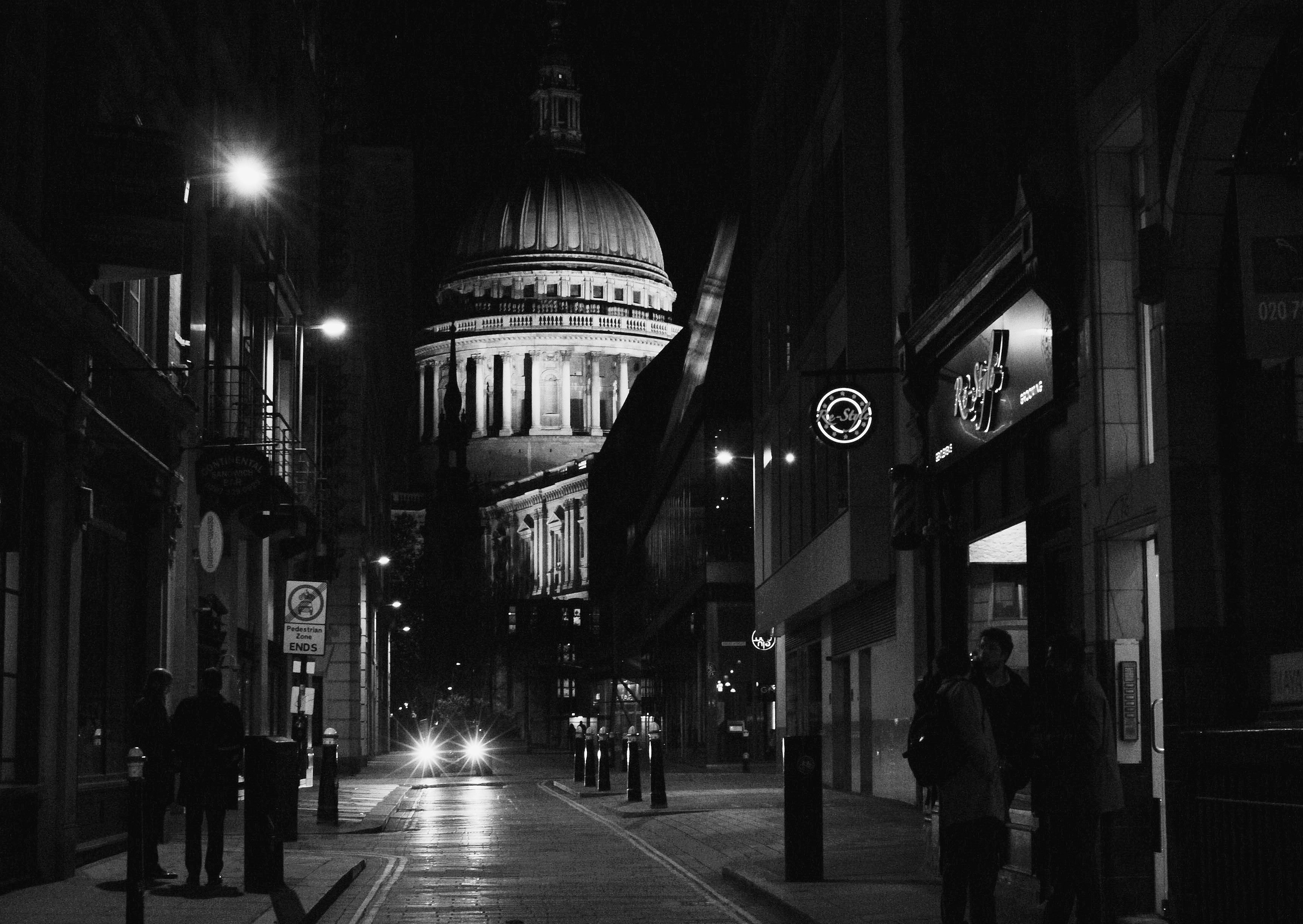 Black and white image looking down a street at night. There are pedestrians on either side of the street and a car headlights shiningr