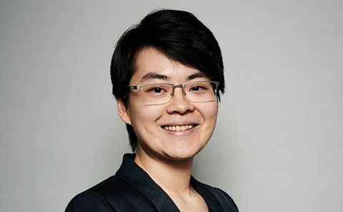 A person with short dark hair, wearing glasses and a dark blazer smiles directly at the camera while posted against a plain light background.