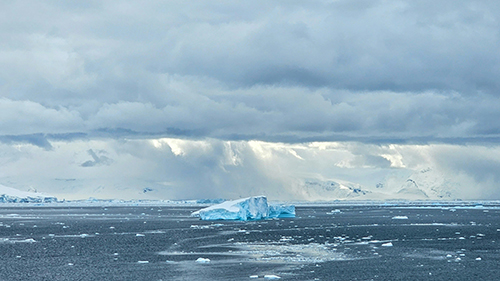 A blue iceberg floats in a dark, icy sea beneath dramatic storm clouds, with snow-covered mountains and falling snow in the distance across a polar landscape.