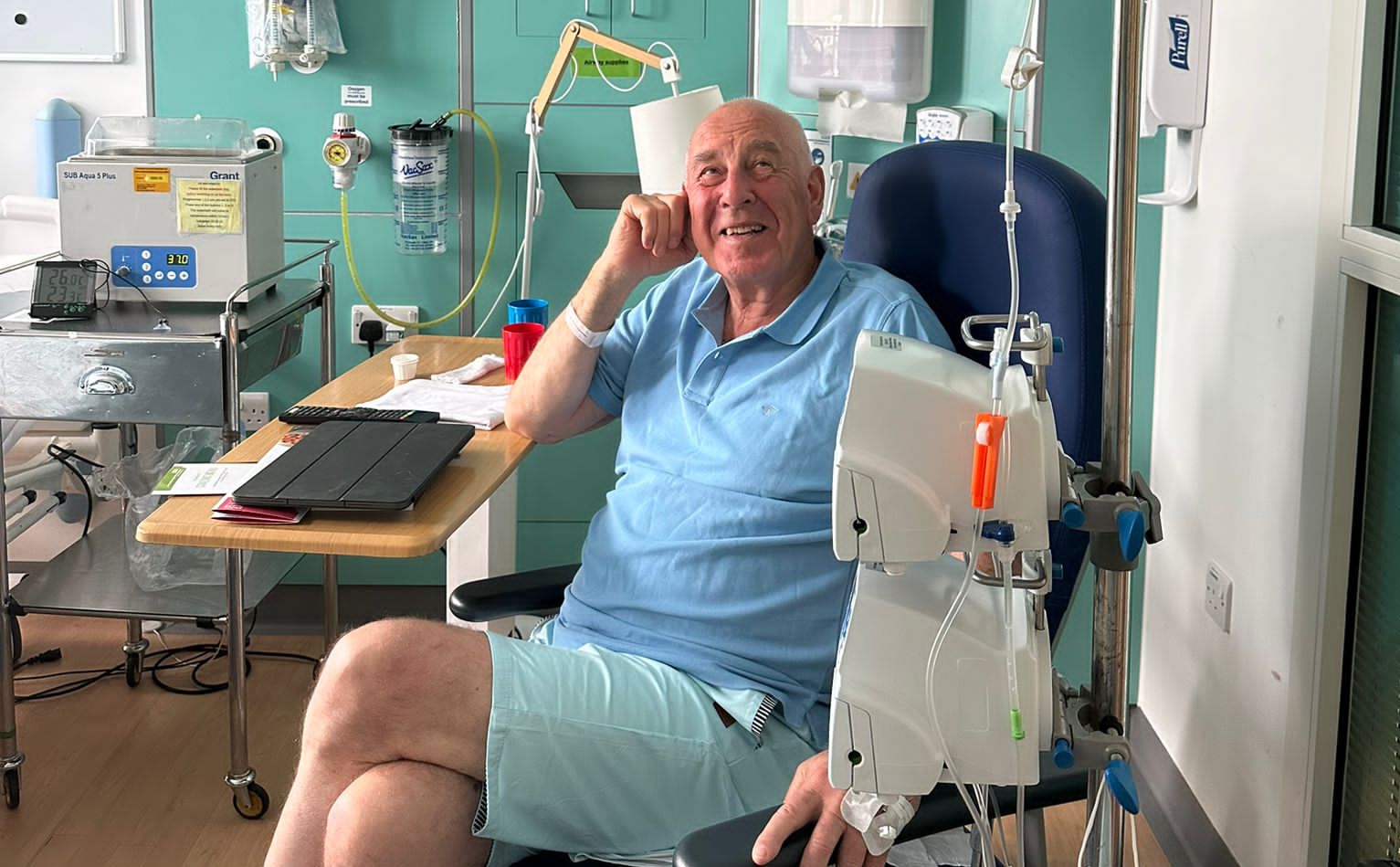 Man in pale blue T-shirt sat in hospital chair, smiling