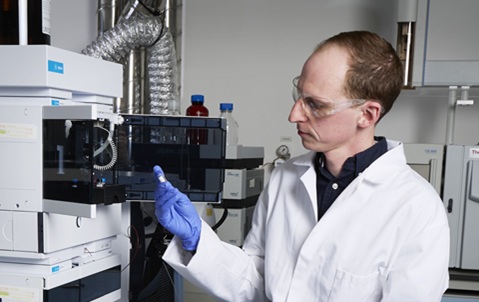 A man wearing a white lab coat, blue gloves and clear goggles sitting in a lab, looking at the contents of a test tube he is holding in his hand