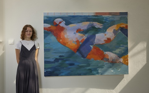 Woman with shoulder length curly hair, wearing a long black dress with a vest top and a white T-shirt underneath, stood next to a large artistic painting of a man swimming in a pool