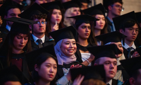 Rows of young people wearing black gowns and black mortar board hats, all looking in one direction. The image is focused on one young woman who is wearing a head scarf under her mortar board and is smiling and clapping