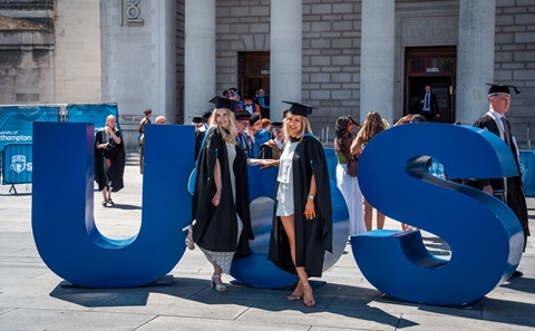 Two young women wearing black gowns and mortar board hats standing in front of giant blue letters spelling UOS