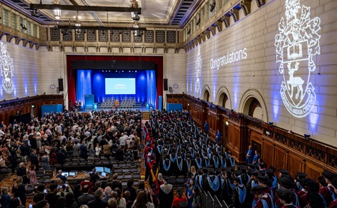 Inside a large guildhall, looking from the back towards the stage. There are dozens of people inside, with those on the right wearing black, blue and red graduation gowns. ’Congratulations’ and the university coat of arms are projected onto the large side wall.