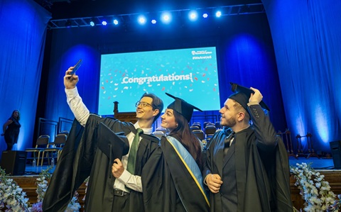 Three people wearing mortar board hats and black graduation gowns look at a mobile phone for a selfie photo 