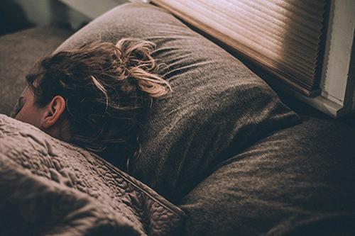Close-up side view of a person sleeping in bed with their head resting on a dark gray pillow next to a window with lowered Venetian blinds. Soft light filters through the blinds, casting warm shadows over the bed. The person has wavy brown hair tied in a messy bun, and a patterned, quilted blanket is pulled up over their shoulder.