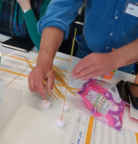 Person assembling a structure with spaghetti and marshmallows on a table.