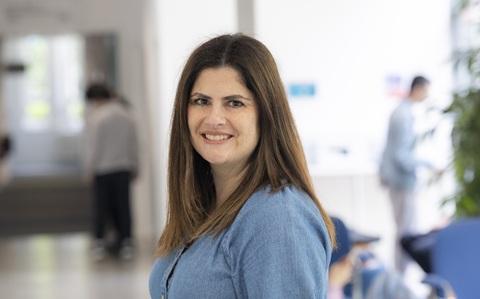 A woman with long straight dark hair, wearing a blue shirt, standing and smiling