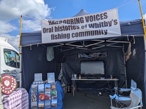A stall with a navy canopy displays a banner reading "Oral histories of Whitby’s fishing community."
