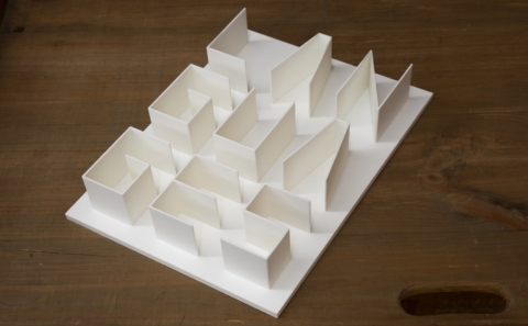 A 3D paper model featuring a grid-like structure of white rectangular walls arranged in varying orientations, displayed on a wooden surface.