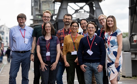 A group of nine people standing, smiling at the camera. They are all wearing red lanyards round their necks. They are stood outside, in front of large bridge structures which are out of focus in the background.