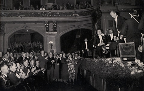 Black and white photo of an audience looking up at an orchestra on stage