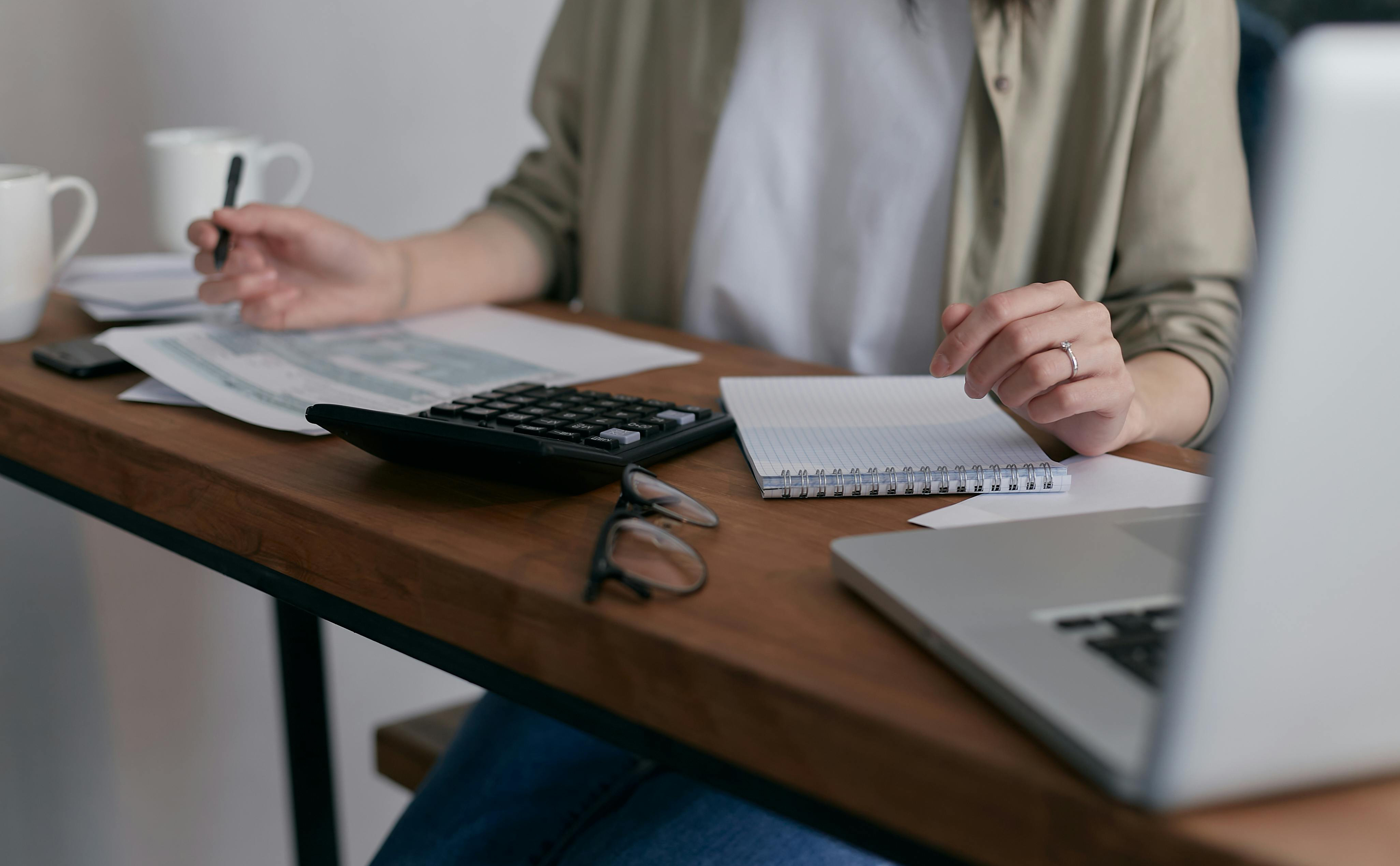 A wooden desk with a woman sat behind it, whose face isn’t visible, holding a pen and working at a laptop.