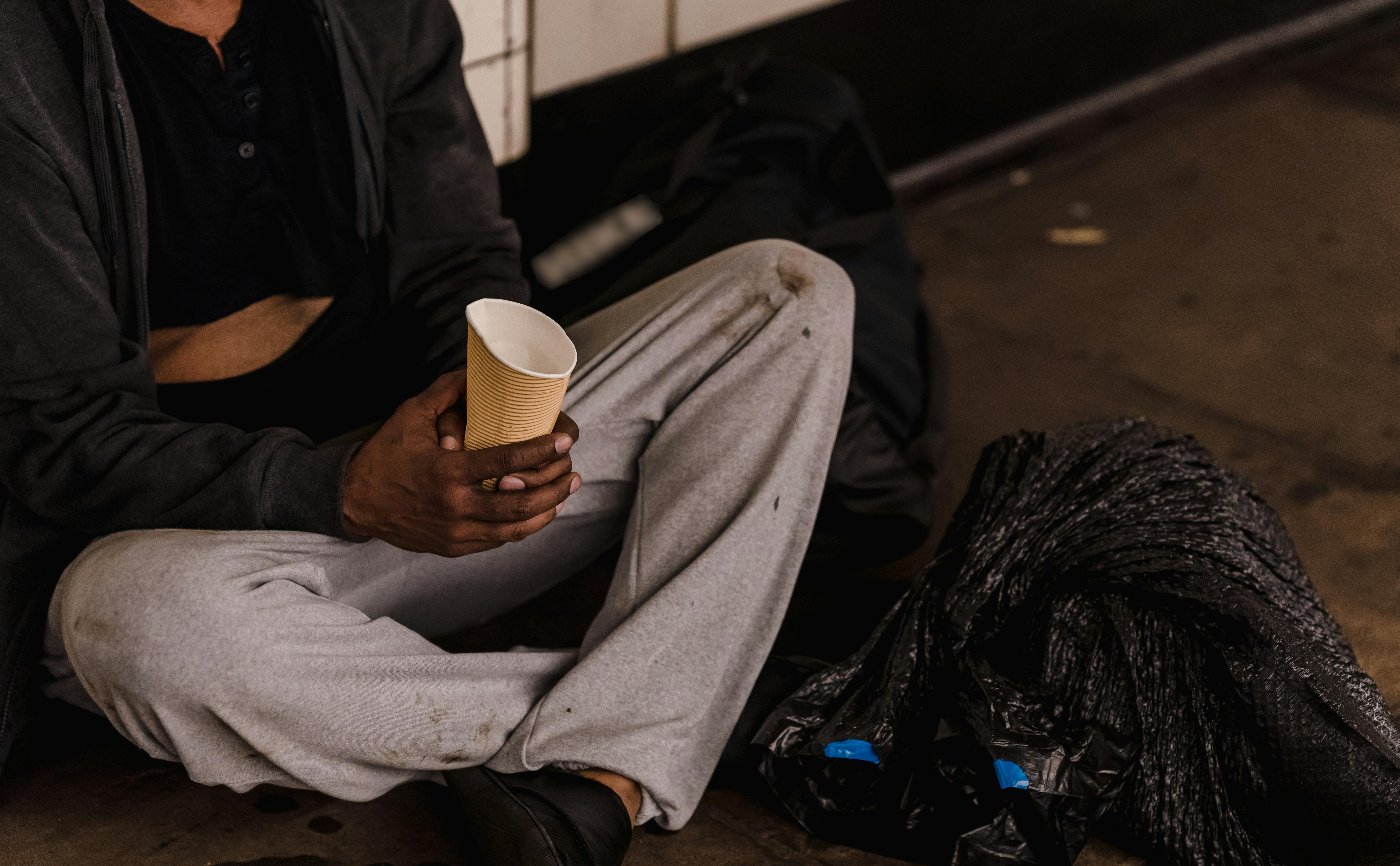 Homeless person shown from the shoulders down sitting on the pavement holding a cup to beg for money, with a black plastic bag in the foreground.