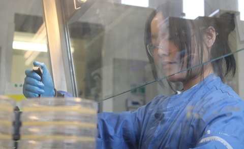 A woman with long hair tied back, wearing a blue lab coat, holds a pipette behind a glass screen