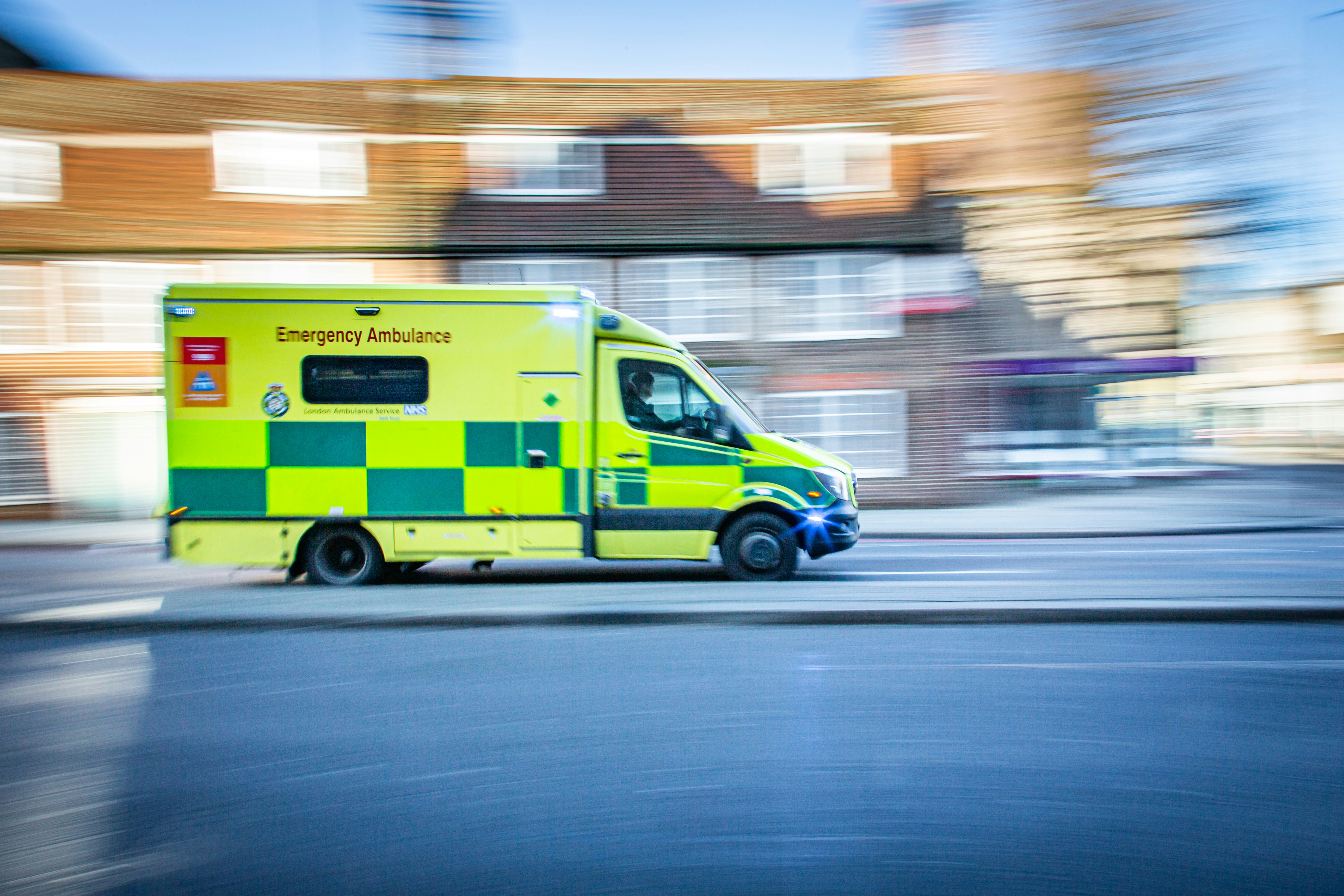 A yellow and green emergency ambulance is speeding down a city street with motion blur in the background, indicating rapid movement. The ambulance is marked with