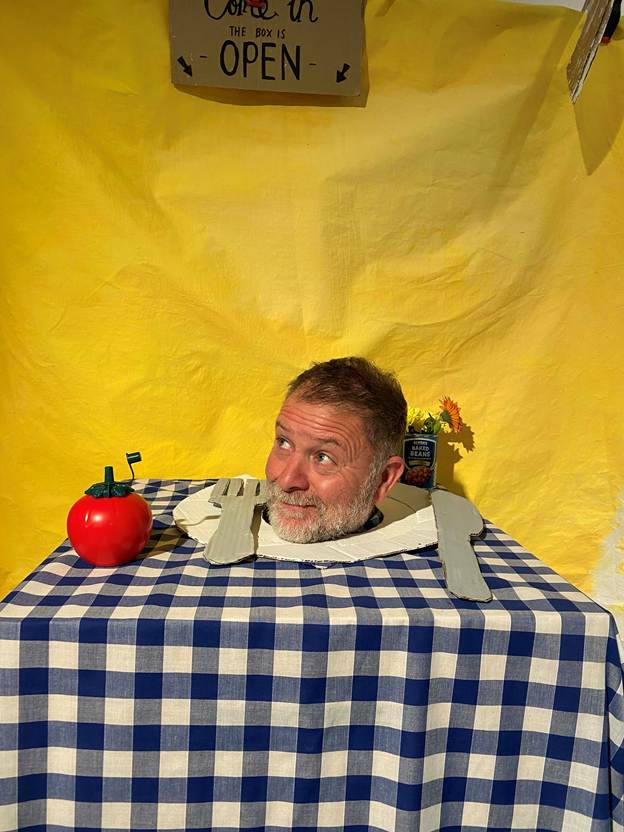 An art installation featuring a table covered with a blue and white check table cloth. There is a plate and tomato-shaped ketchup bottle, and an over-sized knife and fork. A man is standing under the table, with only his head visible on the plate. 