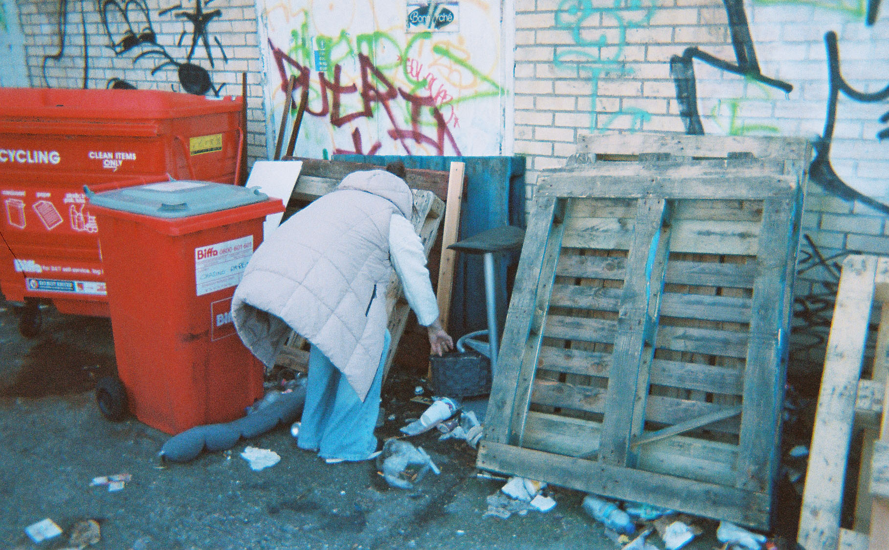 A homeless woman seen from behind, bent over next to bins in an urban area with graffiti on the walls.