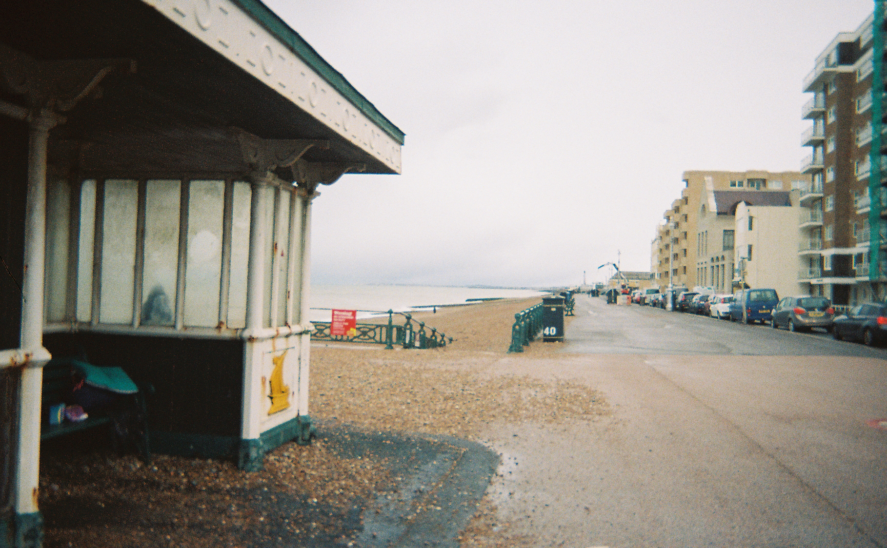 A seafront scene with a street running into the distance in the centre, modern flats on the right side of the road and a traditional seaside shelter on the left in the foreground.