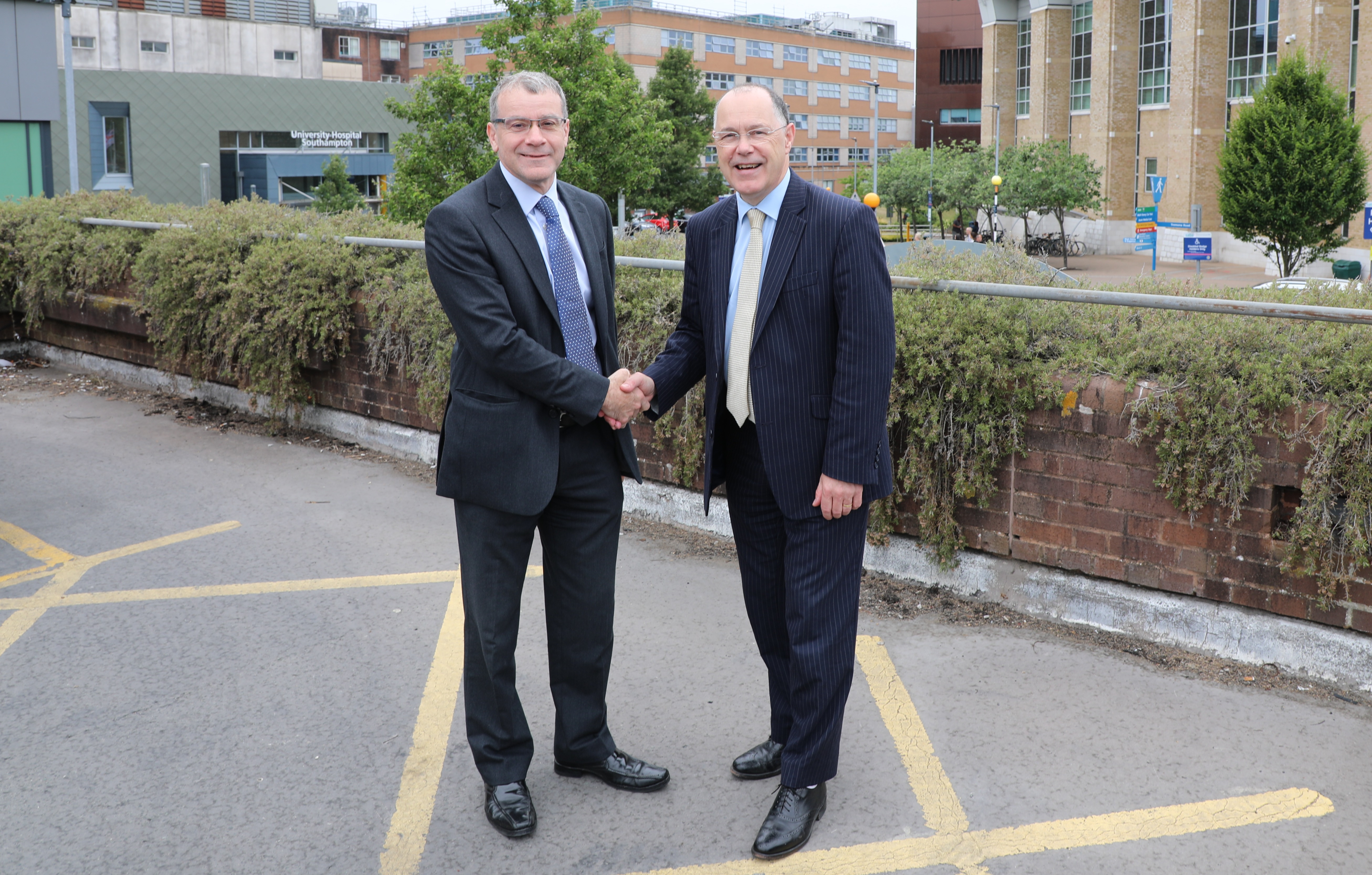 Two smartly dressed men smile and shake hands. Hospital buildings can be seen in the background. 