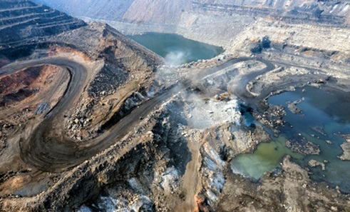 Aerial view of dirt roads winding down hills into a large area that has been dug out and parts of which are filled with water. There is an orange digger in the centre of the image, which looks tiny and indicates the huge scale of the area shown.