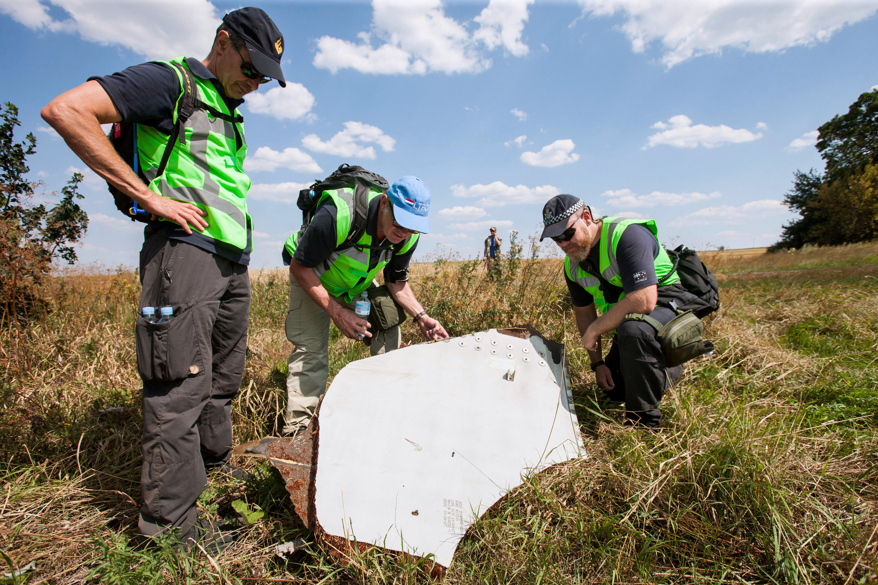 Three men in high-vis tabbards and wearing caps, stood in a grassy field, looking at a large white piece of metal