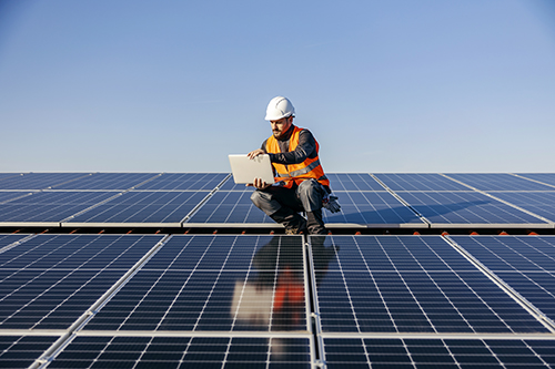 Man in hard hat and high vis standing on a roof top looking at plans. There are solar panels install in front and behind him. 