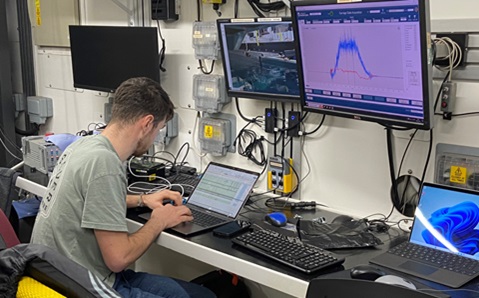 Man in grey T-shirt sat at a desk with multiple laptops, keyboards and monitors in front of him