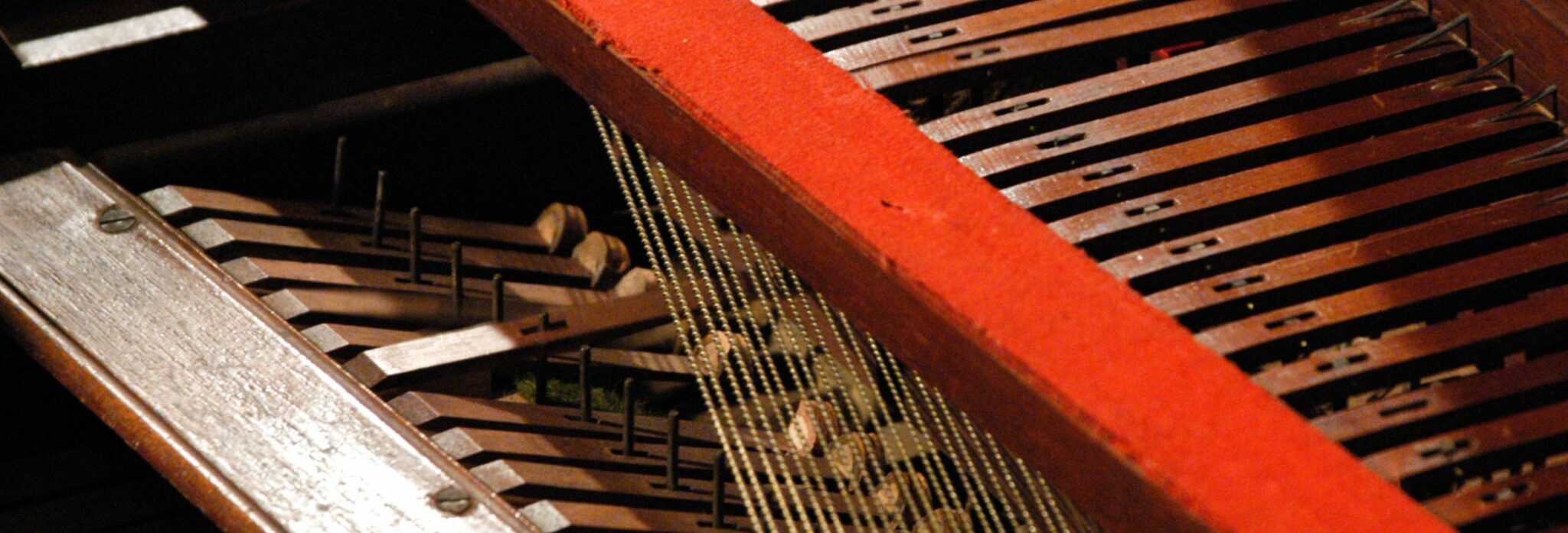  A close-up view of the internal mechanics of a musical instrument, likely a harpsichord or a piano, showcasing wooden levers, strings, and metal pins. The image features a vibrant red felt lining along the edge.