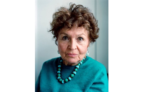 Head and shoulders image of a woman with short curly brown hair looking into the camera. She is wearing a teal coloured jumper and beaded necklace. 