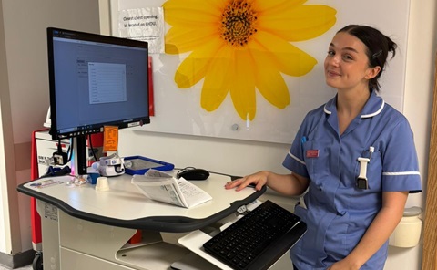Young woman in a blue nurse’s uniform standing beside a computer, with a large yellow flower image on the wall behind