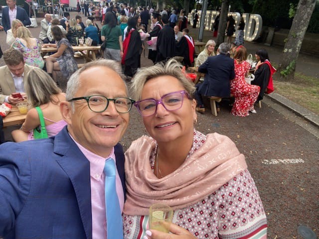 Man and woman, both wearing glasses, smiling up at the camera, stood in front of a crowd of people. 