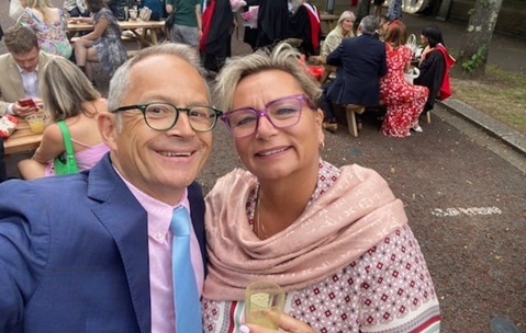 A man in a blue suit, pink shirt and blue tie, and a woman wearing a pink wrap, both smiling at the camera