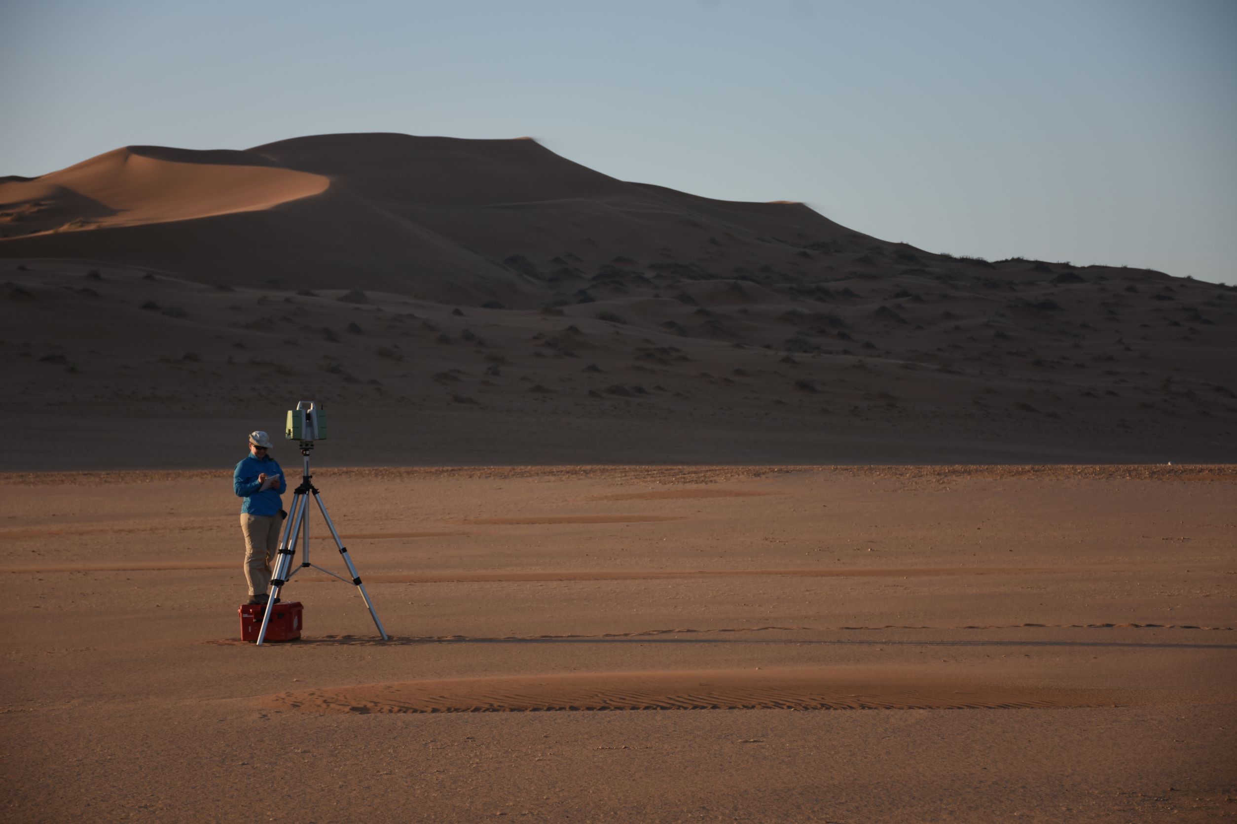 A person conducting research in a desert with dunes in the background, using a surveying instrument on a tripod.