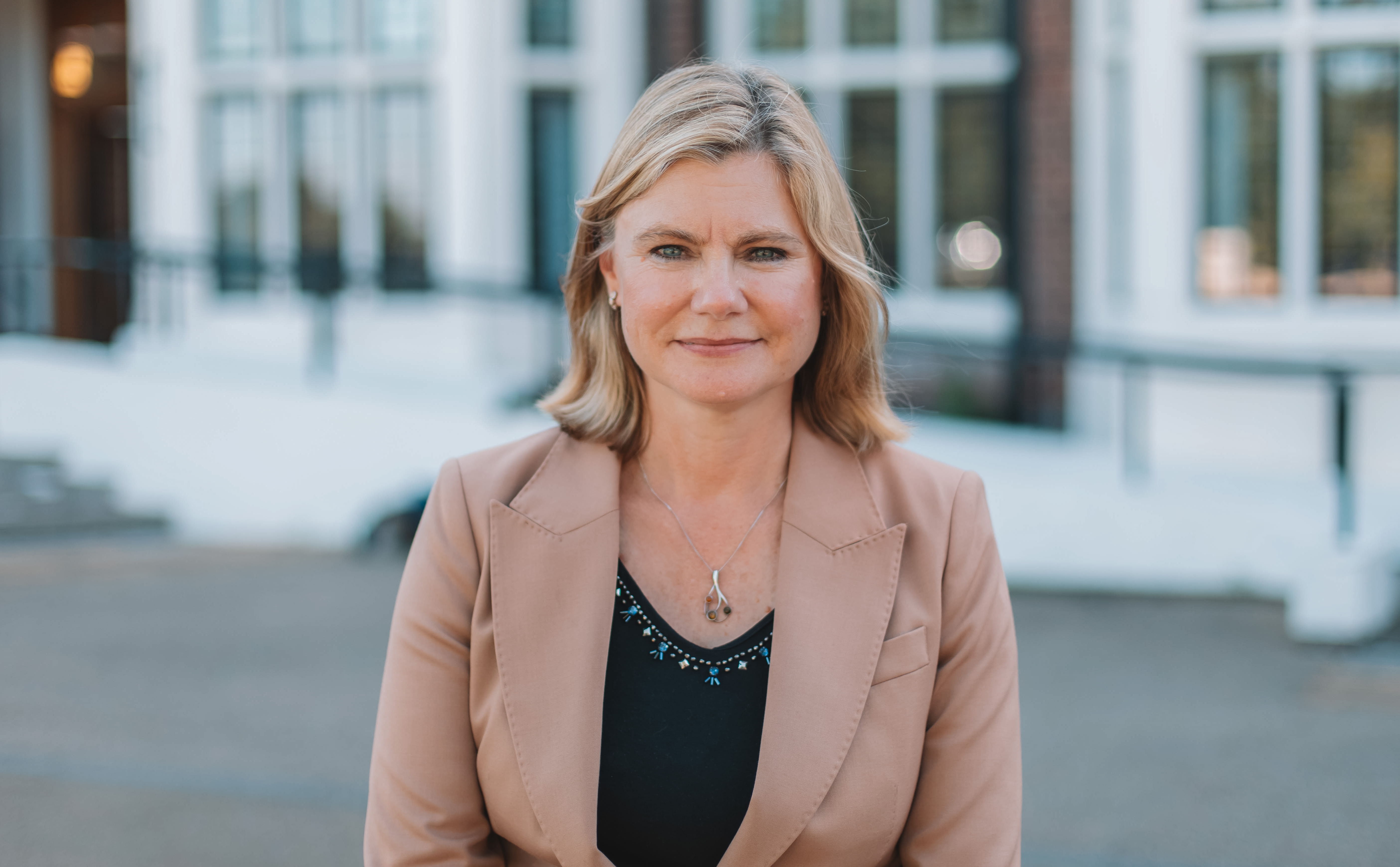 Woman with shoulder length blonde hair, wearing a black top and beige blazer