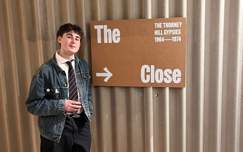 Man wearing a white shirt, tie and denim jacket holding a glass of drink, standing next to a sign reading 'The Close' with an arrow pointing to the right