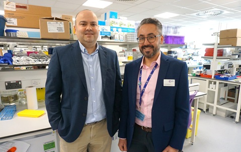 Two men, wearing navy blazers and shirts, standing side by side inside a medical research lab