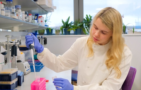 A woman with long blonde hair wearing a white lab coat, sitting at a desk in a lab using a pipette