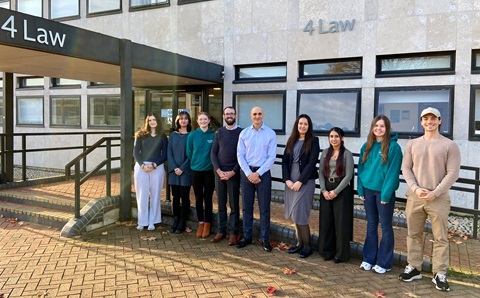 Nine people standing in a line, outside in front of a building. The building has a sign saying ’4 Law’ on the wall. 