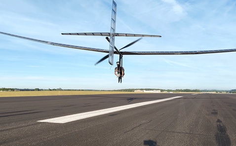 Rear shot of a man sat in an open glider-style plane, flying a few metres above the ground along a runway under a clear blue sky