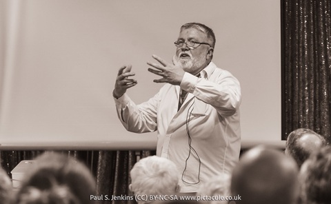 A man with a beard and glasses, wearing a white lab coat, standing in front of an audience talking