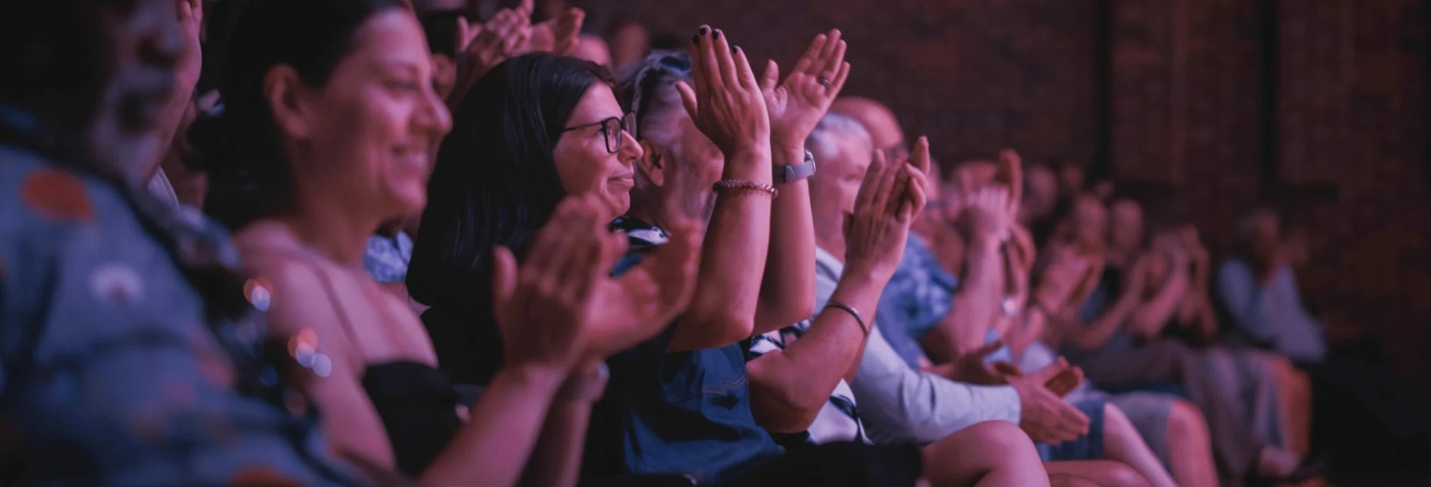 The audience sits in rows clapping toward a stage, with hands raised and illuminated by soft purple lighting. The scene shows a crowded indoor performance venue with people closely seated and applauding enthusiastically.