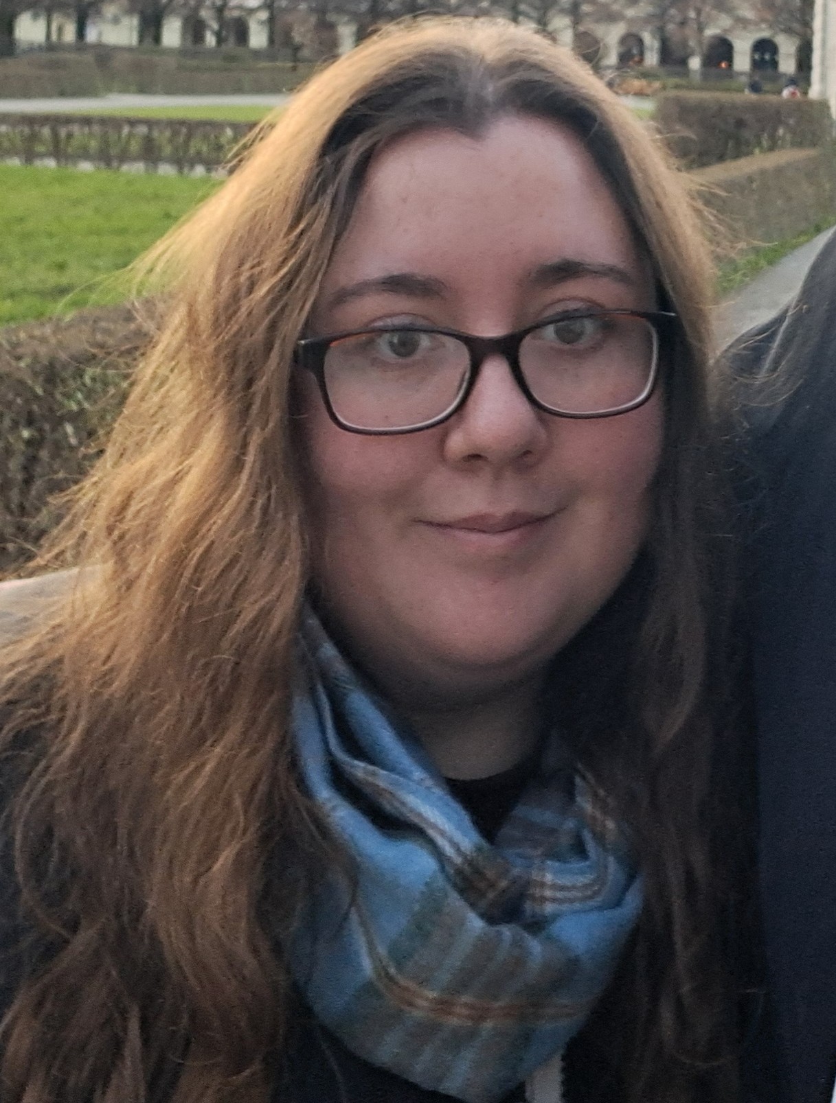 A person with long hair and glasses is shown outdoors wearing a patterned scarf, with hedges and a park landscape in the background. The image is taken at close range with soft daylight.