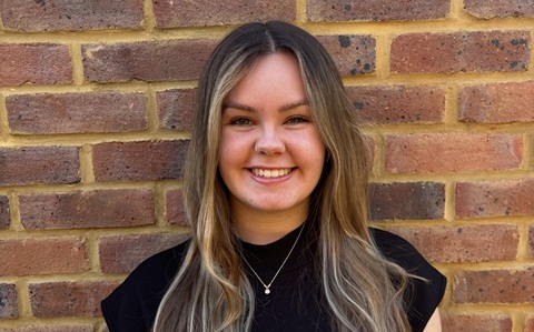 Young woman with long hair smiling at the camera, stood in front of a plain brick wall