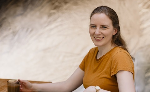 Woman with long brown hair, wearing a yellow T-shirt, smiling at the camera. She is holding a cup of tea and is in front of a light brown blurred background