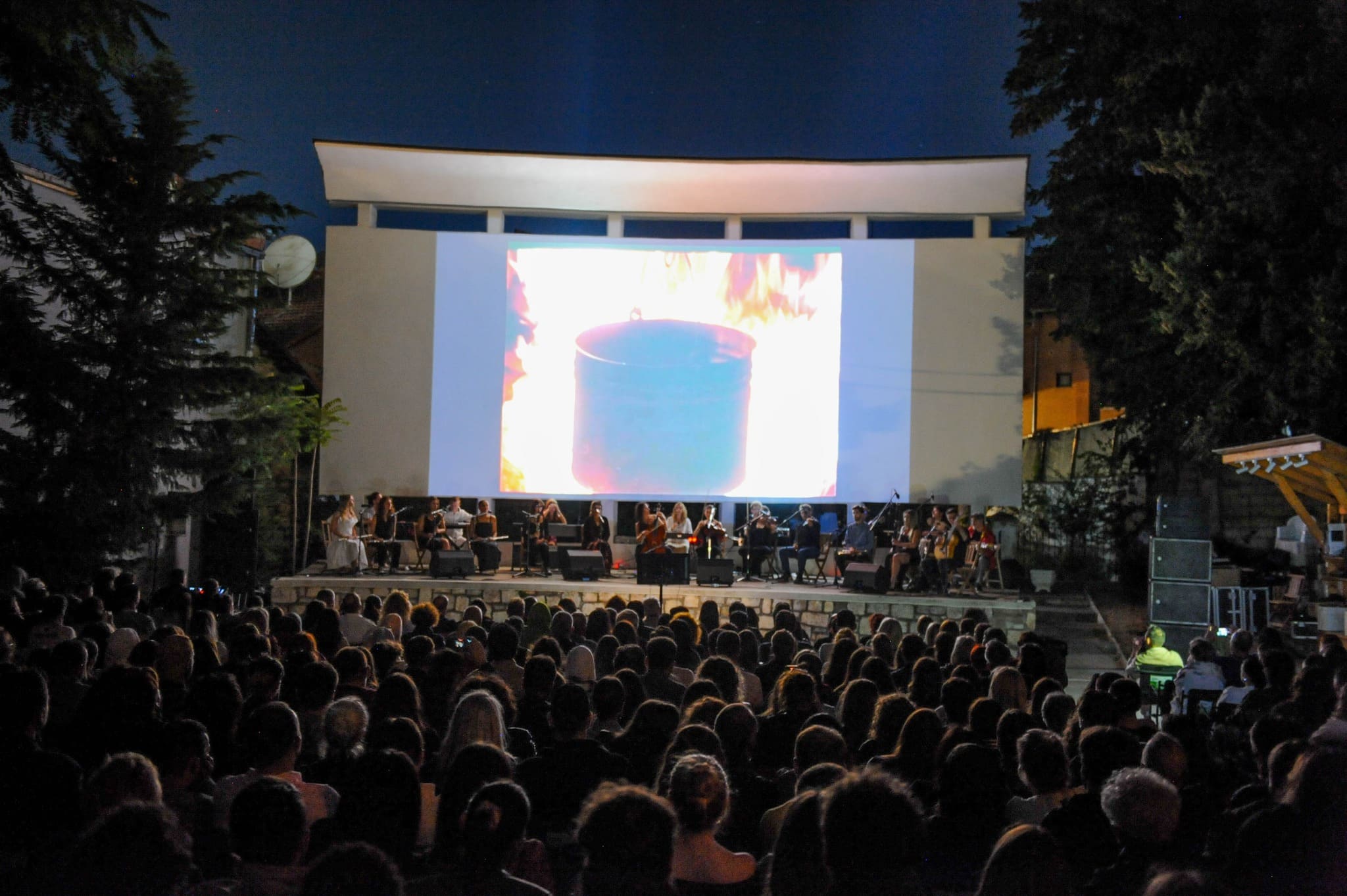 An outdoor nighttime event with a large audience seated in front of a stage featuring musicians and a projection screen displaying an image of a pot over flames. The setting is surrounded by trees and illuminated by stage lights.