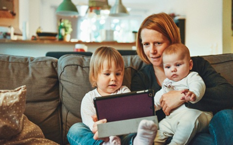 A woman sitting on a sofa with a baby on her lap and a toddler by her side. They are all looking at a tablet screen. 