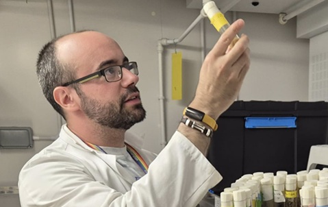 A man wearing glasses and a white lab coat, holding up a test tube and looking at it