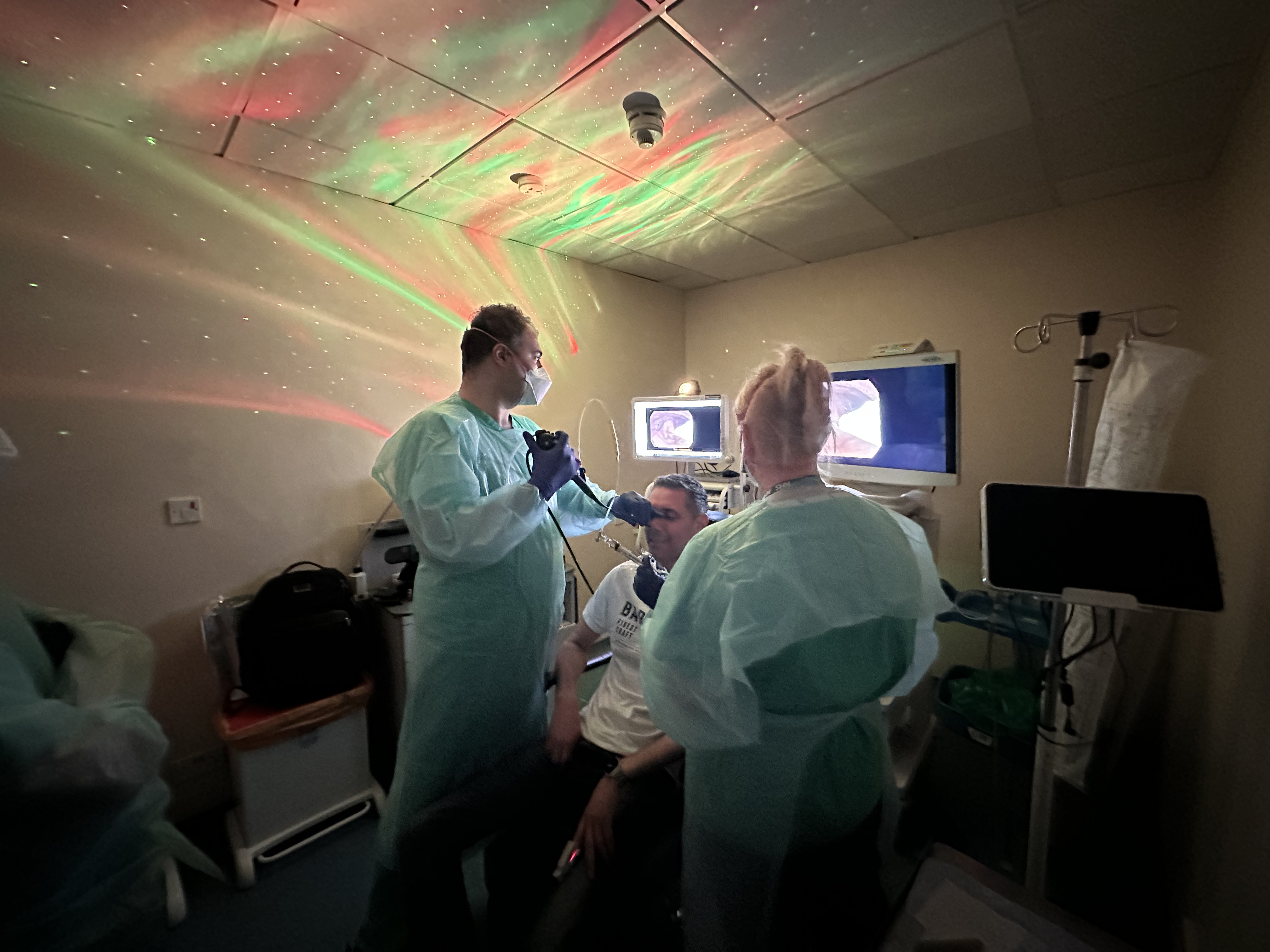 Three healthcare workers in protective gowns and face masks stand around a seated patient in a small clinical room. One clinician appears to be performing a procedure near the patient's face while medical monitors display images in the background. Coloured green and red light patterns are projected across the ceiling and walls.